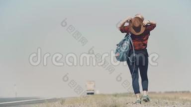 年轻时尚的女孩穿着牛仔衣服，在沙漠里搭便车，在路上。 她背着背包和帽子。 通行证